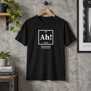 May include: A black short-sleeved t-shirt featuring a white graphic designed like a periodic table element box. The graphic displays the number 123, the text Ah!, the number 230 in brackets, and the phrase The Element of Surprise! below it.
