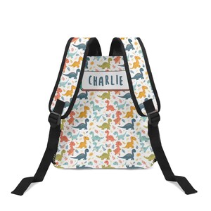 May include: A white backpack featuring a repeating pattern of colorful cartoon dinosaurs and small leaves. The bag has black straps and a rectangular label on the back panel with the name CHARLIE printed in dark blue capital letters.
