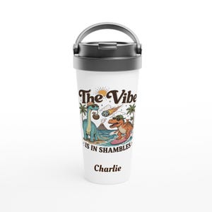 May include: A white travel tumbler with a black lid and handle featuring a graphic of two dinosaurs, a volcano, and a meteor with the text The Vibe Is In Shambles and the name Charlie printed in brown letters on the front of the container.