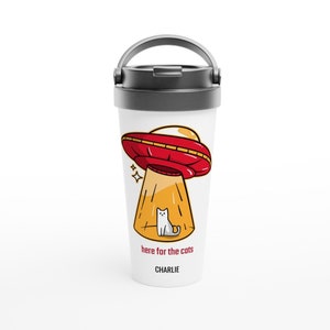 May include: White travel mug featuring a red and yellow UFO design. The UFO is shining a light on a white cat. The text on the mug reads "here for the cats" and "CHARLIE".