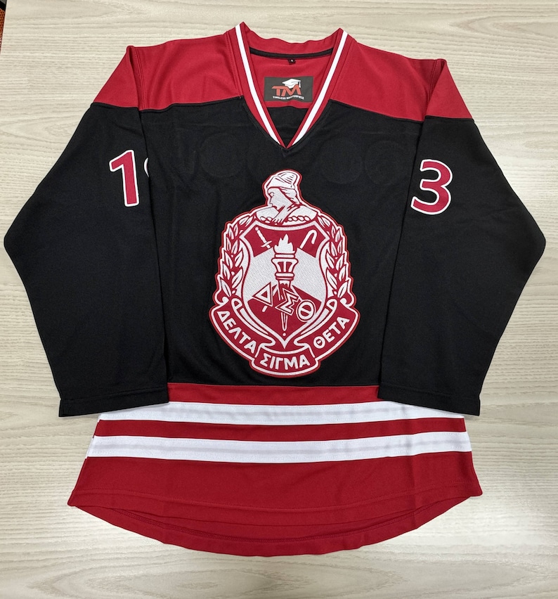 May include: Black and red hockey jersey with a white and red crest featuring a torch and the text "Delta Sigma Theta". The jersey has the numbers "1" and "3" on the sleeves.