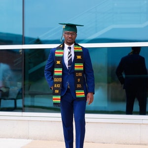 May include: A man wearing a blue suit and a green graduation cap, standing in front of a glass building. He is wearing a black and gold graduation stole with the text "Educated Black Man" and "Class of 2021".