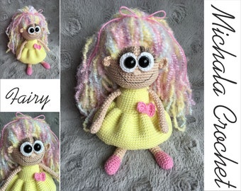 May include: A crocheted fairy doll with long, colorful hair, wearing a yellow dress and pink shoes. The doll is sitting on a gray textured surface. The text "Michaela Crochet" is visible in the background.