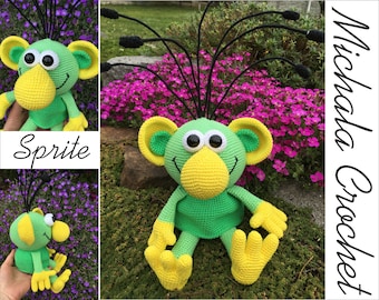 Può includere: Un giocattolo di peluche verde all'uncinetto con un becco giallo e grandi occhi. Il giocattolo ha lunghi tentacoli di filato nero sulla cima della testa. Il giocattolo è seduto su un letto di muschio verde.