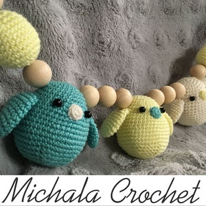 Puede incluir: Una guirnalda de ganchillo hecha a mano con tres adornos en forma de pájaro en verde azulado, amarillo y crema, intercalados con cuentas de madera. Los pájaros tienen ojos negros y detalles blancos. El texto "Michala Crochet" está en la parte inferior.