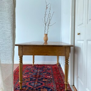 May include: A rustic wooden table with turned legs sits on a red, blue, and cream patterned rug. A vase with dried branches sits on the table.