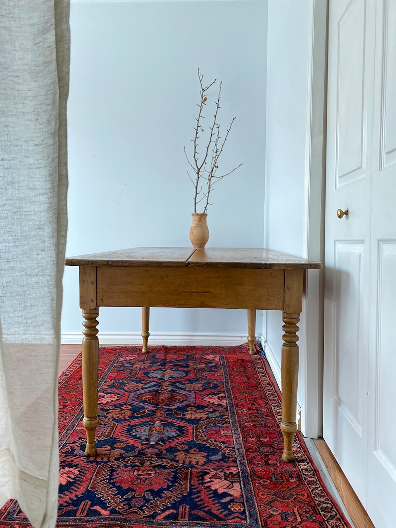 May include: A rustic wooden table with turned legs sits on a red, blue, and cream patterned rug. A vase with dried branches sits on the table.