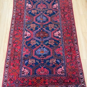 May include: A vintage Persian rug with a red background and a blue central medallion. The rug has a geometric pattern with floral motifs and a fringe border.