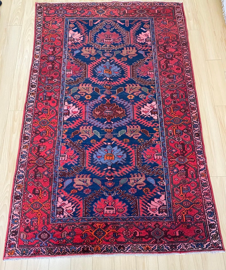 May include: A vintage Persian rug with a red background and a blue central medallion. The rug has a geometric pattern with floral motifs and a fringe border.