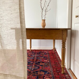 May include: A wooden table with turned legs sits on a red and blue patterned rug. A vase with dried branches sits on the table.