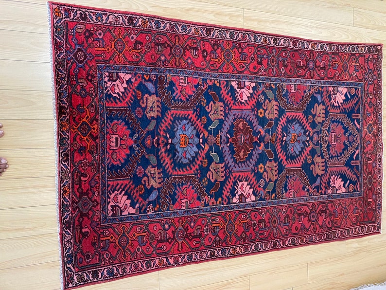 May include: A traditional Persian rug with a deep blue center and a red border. The rug features intricate floral and geometric patterns in shades of pink, red, blue, and purple.