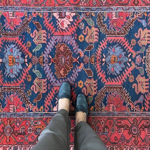 May include: A pair of black leather loafers with a pointed toe are worn by a person standing on a red, blue, and white patterned rug.