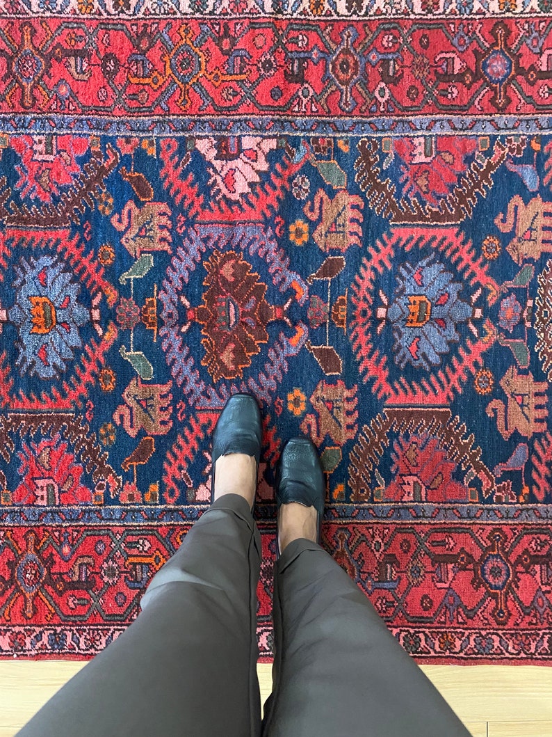 May include: A pair of black leather loafers with a pointed toe are worn by a person standing on a red, blue, and white patterned rug.