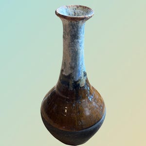 Puede incluir: Un pequeño jarrón de cerámica hecho a mano con una base bulbosa y un cuello delgado. El jarrón presenta un acabado marrón brillante en la mitad inferior, que transiciona a un gris y blanco más claro y texturizado en la parte superior. El jarrón mide unos 15 cm de alto.