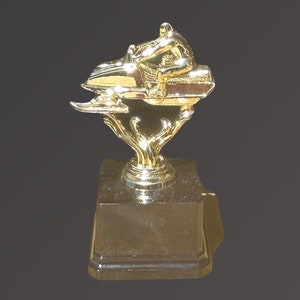 May include: A gold-colored trophy with a figure of a person riding a snowmobile. The trophy has a square base.