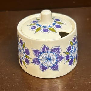May include: A small, round, ceramic sugar bowl with a lid. The bowl and lid are white with a repeating floral pattern in blue and purple. The lid has a small handle and a spoon slot.