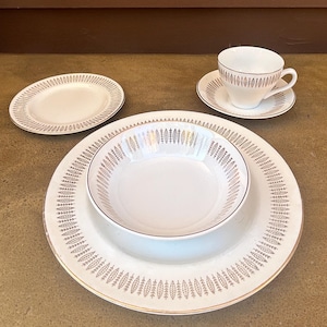 Puede incluir: Un juego de vajilla de porcelana blanca con detalles de pan de oro. El juego incluye un plato llano, un cuenco, un platillo, una taza de té y un plato pequeño. Los platos están dispuestos sobre una superficie marrón.