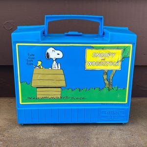 May include: A blue plastic lunchbox featuring Snoopy and Woodstock. The front panel has an illustration of Snoopy sitting on his doghouse with Woodstock. The words "Snoopy and Woodstock" are in a yellow rectangle. The lunchbox has a handle.