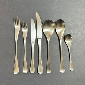 May include: A set of seven silver-toned cutlery pieces, including two forks, two knives, and three spoons. The cutlery is arranged in a row on a dark grey surface.