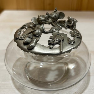 Könnte beinhalten: Eine klare Glasschale mit einem runden Zinndeckel. Der Deckel ist mit skulpturalen Figuren von Mickey und Minnie Mouse sowie kleinen Schmetterlings- und Blumenmotiven verziert. Der Metalldeckel liegt fest auf dem Rand der Glasschale auf.