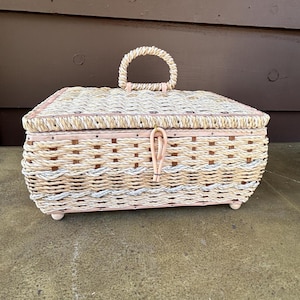 May include: A woven wicker storage box with a pink painted finish and a rope handle. The box has a woven pattern and is sitting on a concrete surface.