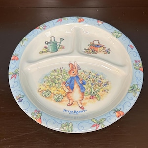 Puede incluir: Un plato blanco dividido con un borde azul y un diseño de Peter Rabbit. El plato presenta una regadera azul y blanca, una cesta de bayas y Peter Rabbit sosteniendo una regadera en un jardín. El texto "Peter Rabbit" está impreso en el plato.
