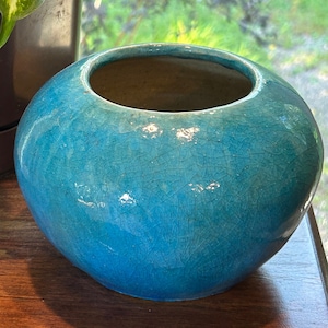 May include: A turquoise blue ceramic planter with a crackled glaze finish. The planter is round and has a wide opening.