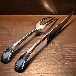 Pode incluir: Um conjunto de talheres para trinchar prateado, incluindo um garfo e uma faca, repousa sobre uma superfície de madeira escura. As pegas têm uma forma arredondada em forma de gota e as lâminas são polidas. O conjunto é projetado para trinchar e servir carnes.