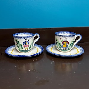May include: Two hand-painted ceramic teacups and saucers. The cups and saucers have a white base with blue rims and yellow accents. Each cup features a different folk art figure. The cups have curved handles.