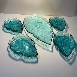 May include: A set of five teal-coloured glass dishes shaped like leaves. The dishes feature a textured surface that mimics the veins of a leaf. One large central dish is surrounded by four smaller dishes, all with a translucent finish.
