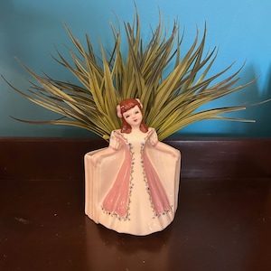 May include: A vintage ceramic planter shaped like a woman in a long, pale pink dress with pink and purple floral details. The planter holds green and yellow spiky leaves. The woman has brown hair and a pink bow.