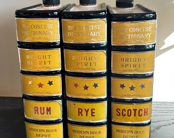 Puede incluir: Un mueble licorero decorativo de cerámica con forma de libros apilados. Cada libro tiene una etiqueta amarilla con texto negro. Las etiquetas dicen "The Concise Dictionary", "Bright Spirit" y los nombres de licores "Rum", "Rye" y "Scotch".