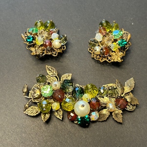 Puede incluir: Un conjunto de tres broches de tono dorado con piedras de vidrio verde, amarillo y rojo. Los broches tienen forma de hojas y flores.