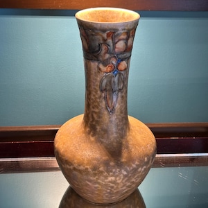 May include: A decorative ceramic vase with a bulbous base and a slender neck. The vase features a mottled brown and tan color scheme with floral and geometric patterns in shades of brown, blue, and orange. The vase is approximately 10 inches tall.