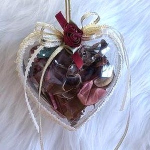 Könnte beinhalten: Herzförmiger Ornament mit getrockneten Blumen und Potpourri gefüllt. Das transparente Kunststoffherz ist mit Spitze und einem goldenen Band verziert und wird von einer goldenen Schnur gehalten. Eine rote Rose und eine Schleife schmücken die Oberseite.