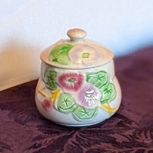 May include: A small, white ceramic jar with a lid, decorated with pink and green floral designs. The jar has a rounded shape and a smooth, glossy finish.