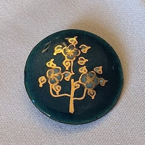May include: A round, dark green enamel pin featuring a gold tree design. The tree has small, gold flowers and leaves.