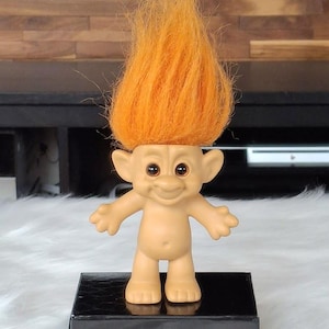 Puede incluir: Una muñeca troll vintage con pelo naranja. La muñeca tiene una cara sonriente, orejas pequeñas y brazos extendidos. Está de pie sobre una base rectangular negra, sobre una superficie blanca y esponjosa.