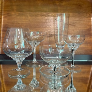 Puede incluir: Un juego de cinco vasos de beber de vidrio transparente, que incluye una copa de champán, una copa de vino y tres copas de coñac. Los vasos están dispuestos sobre una superficie de madera.