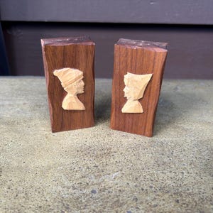 May include: Two wooden carvings of profiles, likely salt and pepper shakers. Each is a rectangular block of dark brown wood with a carved light-colored profile. One wears a hat, the other a head covering. The background is a mottled, light brown surface.