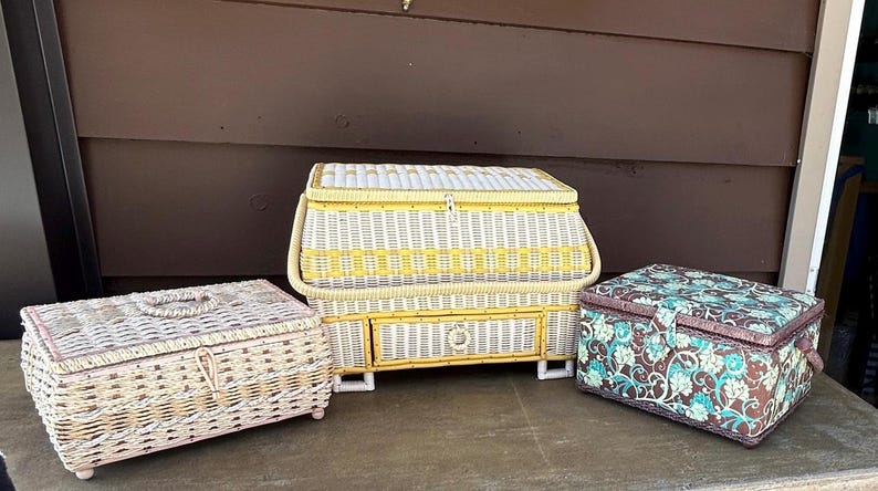 May include: Three vintage wicker sewing boxes. One is white with yellow accents and a drawer. Another is white with pink accents. The third is brown with a floral pattern.