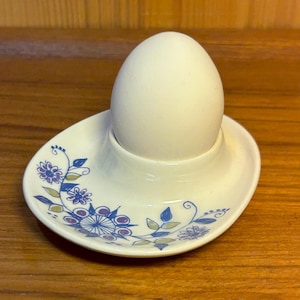 Może przedstawiać: Biały ceramiczny kieliszek do jajek z kwiatowym wzorem w kolorze niebieskim, fioletowym i zielonym, trzymający białe jajko. Kieliszek do jajek ma owalną podstawę i centralny kubek do trzymania jajka. Wzór zawiera stylizowane kwiaty i liście.