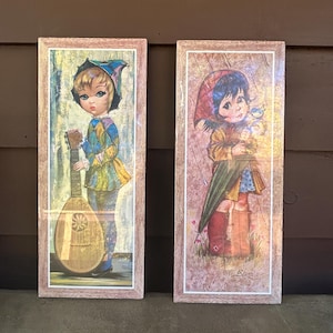 May include: Two framed vintage prints. One depicts a child in a harlequin outfit holding a lute. The other shows a child in a red hood holding an umbrella and a bird. Both prints are in light brown wooden frames.