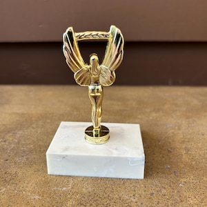 Peut inclure: Un trophée doré représentant une figure ailée sur un socle rectangulaire en marbre. La figure a des ailes déployées et tient une couronne de laurier. Le socle est blanc avec des veines grises.