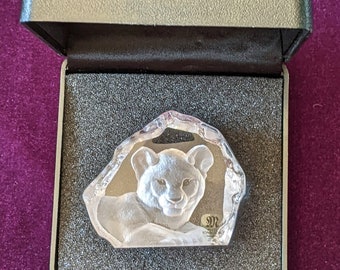 May include: A clear glass paperweight in the shape of a rock with a detailed carving of a lion cub inside. The paperweight is in a black velvet box with a silver latch.