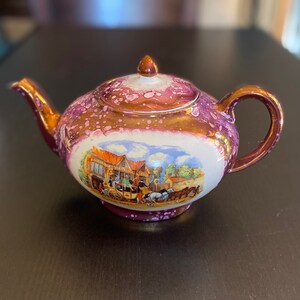 Peut inclure: Une petite théière rose et dorée avec une scène blanche d'une calèche tirée par des chevaux et un bâtiment. La théière a un couvercle et une poignée.