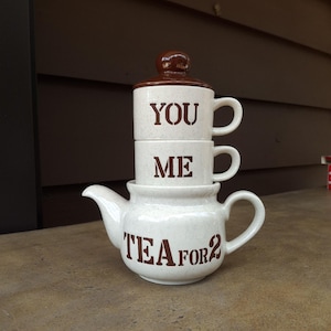 Könnte beinhalten: Eine weiße Keramik-Teekanne mit braunem Deckel und dem Schriftzug "TEA FOR 2" in braunen Buchstaben. Zwei gestapelte Tassen lesen "YOU" und "ME". Die Teekanne und die Tassen sind gesprenkelt.
