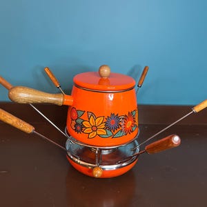 May include: Orange fondue pot with floral design and six wooden fondue forks. The pot is sitting on a metal stand with a glass insert.