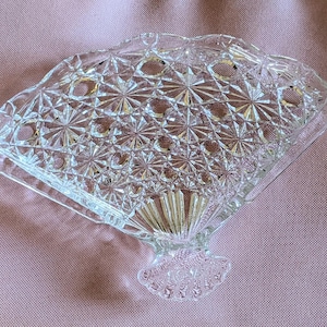 Könnte beinhalten: Ein durchsichtiger Serviettenhalter aus Glas mit einem fächerförmigen Design und einem Diamantenmuster. Der Halter hat einen gewellten Rand und eine dekorative Basis.