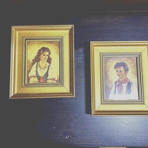 May include: Two framed portraits, one of a girl with long brown hair wearing a white dress with a red sash, and the other of a boy with short brown hair wearing a white shirt with a red scarf.